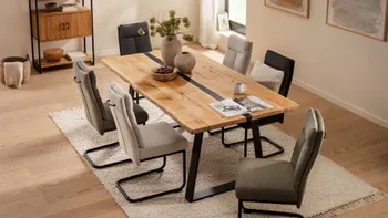 Modernes gemütliches Esszimmer mit einem großen Massivholzesstisch aus Wildeiche, der in der Mitte einen schmalen schwarzen Streifen aus Epoxidharz hat, um den Tisch stehen Freischwinger Stühle in grün, beige und grau.
