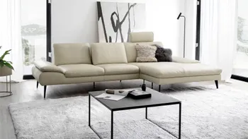 Beiges Ledersofa, bei dem eine Kopfstütze hochgeklappt ist in einem hellen Raum mit großen Fenstern.