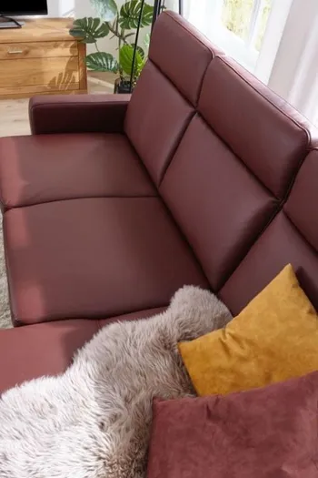 Weinrotes Ledersofa mit hoher Rückenlehne und gelbem und rotem Kissen sowie einem rosa Fell auf dem Sitz.