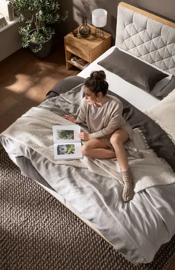 Frau sitzt entspannt auf einem schönen natürlichen Bett mit gestepptem Polster Kopfteil vor ihr ein aufgeschlagenes Buch und blickt in die Ferne.