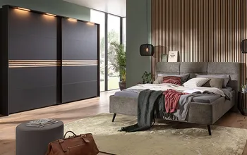 Modernes Schlafzimmer mit grauem Polsterbett und dunklem Schwebetürenschrank mit Holzdekor-Elementen, akzentuiert durch eine Holzlamellenwand, warmes Lichtkonzept und gemütliche Textilien für eine stilvolle und einladende Atmosphäre.