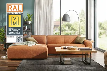 Gütesiegel &quot;Goldene M&quot; auf einem Bild von einem Ecksofa mit Lederbezug in Cognac in einem hellen Wohnzimmer mit dunklem Teppich und Holzboden.