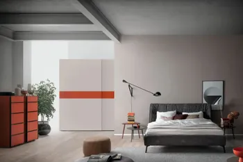 Graues Polsterbett mit schwarzen Füßen und dekoriert mit mehreren Kissen und Decken in Beigetönen in einem modernen Schlafzimmer mit einem großen weißen Schwebetürenschrank mit orangenem Akzent.