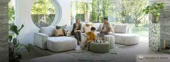 Familie im hellen Wohnzimmer auf heller Couch mit grünem Hocker umgeben von großen Fenstern und Zimmerpflanzen