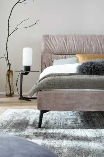 Hälfte eines beigen Polsterbetts mit weißem. gelbem und schwarzem Kissen und einer weißen und grünen Decke in einem hellen modernen Schlafzimmer.