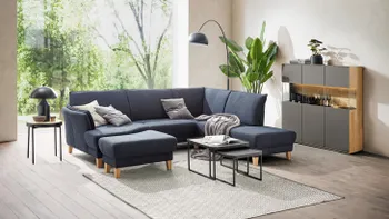 Blaues Ecksofa mit dicken Beinen aus Holz in einem gemütlich eingerichteten Wohnzimmer mit großem Fenster hinter dem Sofa.
