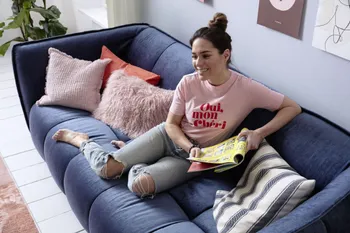Lächelnde Frau sitzt gemütlich mit einer Zeitung in der Hand auf einem blauen Sofa mit Steppung und abgerundeten Ecken und dekoriert mit rosa Kissen.