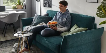 Petrolfarbenes Sofa mit leicht nach oben geklappter Armlehne, auf dem ein Junge sitzt in einem hellen Raum.