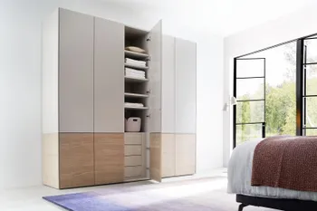 Moderner weißer Kleiderschrank, der am unteren Teil Holzelemente hat und fünf Türen hat, die dritte Türe ist geöffnet und man sieht Regalböden im oberen Teil und Schubladen im unteren Teil.