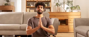 Mann sitzt mit geschlossenen Augen in Buddha Pose in einem hellen Wohnzimmer.